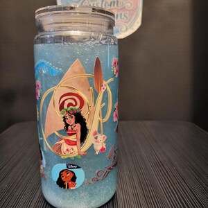 Puede incluir: Un vaso de vidrio transparente con tapa, lleno de l&iacute;quido azul y purpurina. El vaso est&aacute; decorado con im&aacute;genes de Moana y otros personajes de la pel&iacute;cula de Disney. El texto "Ashley's Beautiful Customs" es visible en una pegatina.