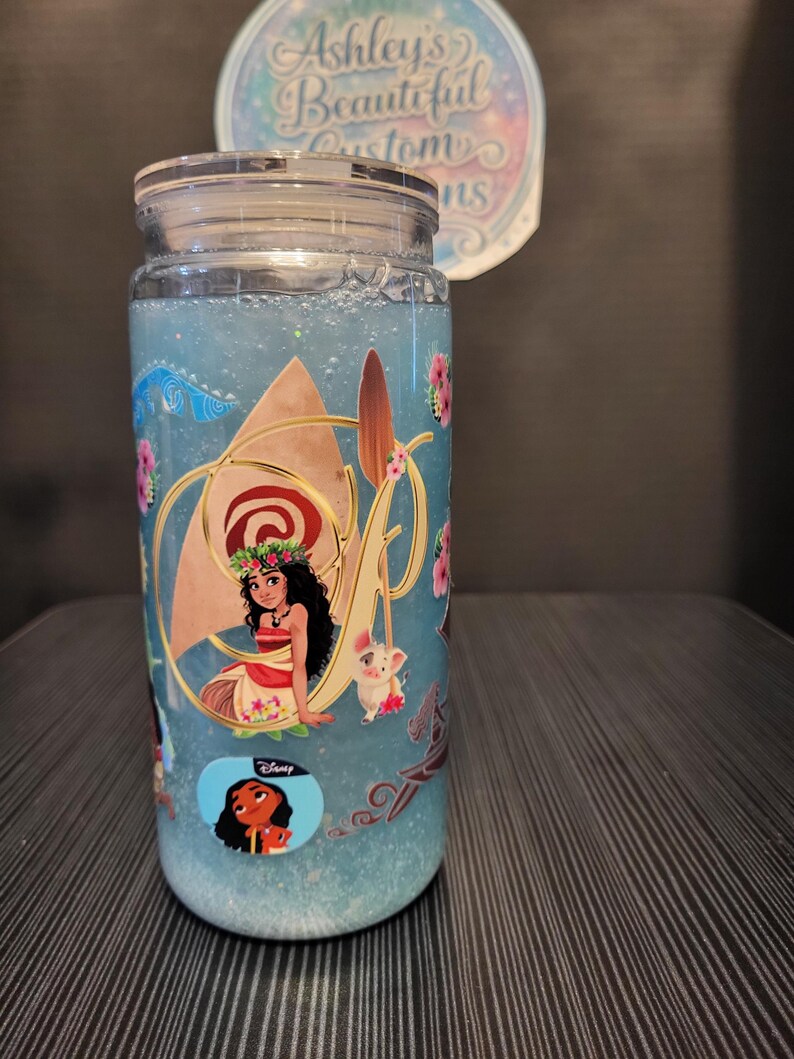 Puede incluir: Un vaso de vidrio transparente con tapa, lleno de l&iacute;quido azul y purpurina. El vaso est&aacute; decorado con im&aacute;genes de Moana y otros personajes de la pel&iacute;cula de Disney. El texto "Ashley's Beautiful Customs" es visible en una pegatina.
