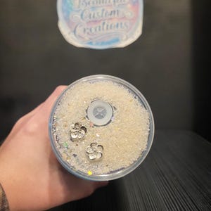 Puede incluir: Un vaso transparente lleno de purpurina blanca y dos abalorios de flores plateadas. El vaso tiene una tapa transparente con un agujero para la pajita. La parte superior del vaso tiene el texto "Ashleys Beautiful Custom Creations".