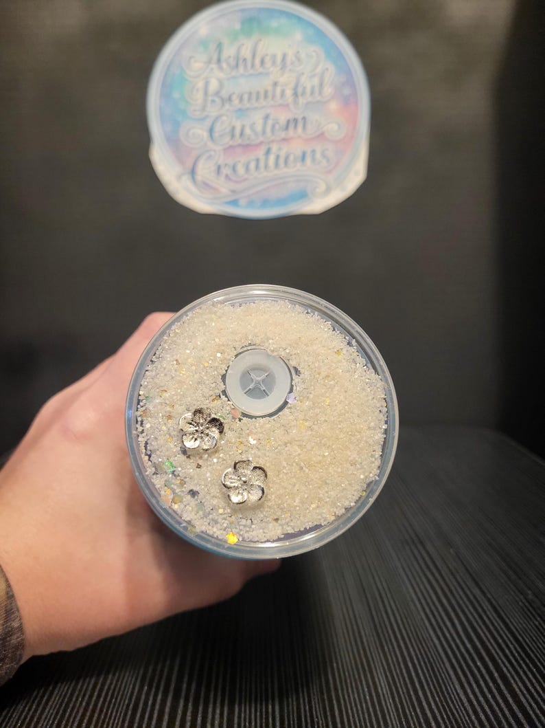 Puede incluir: Un vaso transparente lleno de purpurina blanca y dos abalorios de flores plateadas. El vaso tiene una tapa transparente con un agujero para la pajita. La parte superior del vaso tiene el texto "Ashleys Beautiful Custom Creations".