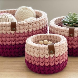 May include: Set of three handmade crochet baskets in varying sizes, featuring a gradient of pink and magenta hues. The baskets have a round shape with a leather strap detail. The largest basket contains yarn, the medium one holds a succulent, and the smallest has wooden beads.