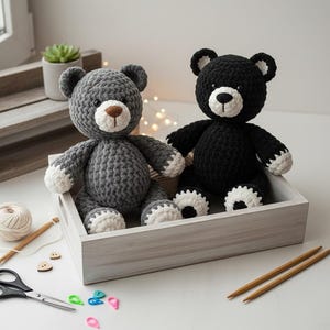 May include: Two handmade crochet teddy bears, one gray and one black, sit in a wooden tray. The gray bear has a brown nose and white accents on its paws and ears. The black bear has white accents. Crafting supplies, including scissors and yarn, are scattered around.