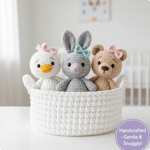 May include: A white crocheted basket contains three handmade soft toys: a duck, a bunny, and a bear. The duck is white with a yellow beak and a pink bow. The bunny is grey with a blue bow, and the bear is tan with a pink bow. The basket has a handle.