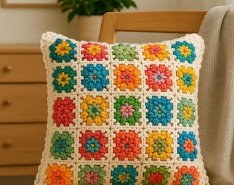 May include: A square crochet pillow with a colorful floral pattern. The pillow features a grid of squares, each with a different colored flower design. The flowers are in shades of red, orange, yellow, green, and blue, with a cream-colored border.
