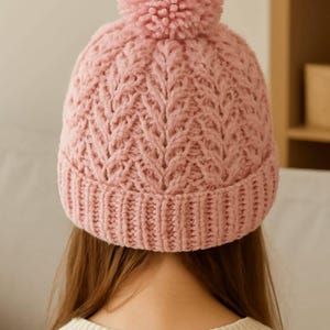 Può includere: Un berretto rosa lavorato a maglia con un motivo strutturato e un pompon abbinato. Il cappello presenta un bordo a coste e un design a trecce dettagliato, perfetto per l'inverno.