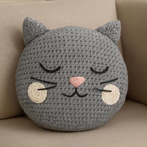 Puede incluir: Un cojín gris tejido a ganchillo con forma de gato, con ojos cerrados, una nariz rosa y mejillas blancas. El cojín tiene orejas puntiagudas y detalles negros bordados para la boca y los bigotes. El cojín está sobre una superficie beige.