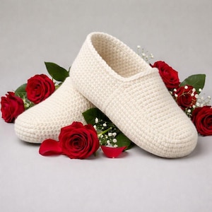Peut inclure: Une paire de chaussons en crochet blanc cassé. Les chaussons sont entourés de roses rouges et de feuilles vertes. Les chaussons ont un design simple à enfiler et une surface texturée. Le fond est gris clair.