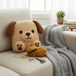 Puede incluir: Un cojín de perro de ganchillo beige hecho a mano con orejas y huellas marrones. Una madeja de hilo amarillo mostaza, un ganchillo y unas tijeras pequeñas están colocadas junto al cojín en un sofá beige. Una manta gris está sobre el brazo del sofá.
