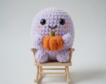 May include: A lavender crocheted creature with black eyes and rosy cheeks, holding an orange pumpkin. The plush toy is seated on a small wooden rocking chair, creating a whimsical and seasonal display.