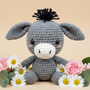 May include: A handmade gray crochet donkey with a white muzzle and black mane, surrounded by pink roses and white daisies. The donkey has large, expressive eyes and is sitting on a neutral background.