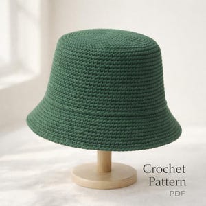 May include: A forest green crochet bucket hat displayed on a wooden stand. The hat has a rounded crown and a slightly downward-sloping brim. The text "Crochet Pattern PDF" is visible in the lower right corner.