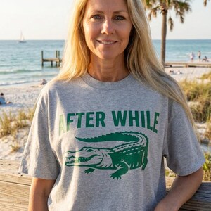 Può includere: Una donna indossa una maglietta grigia con la scritta "AFTER WHILE" e una grafica di un alligatore verde. Indossa jeans blu ed è in piedi vicino a una staccionata di legno con una spiaggia e l'oceano sullo sfondo.