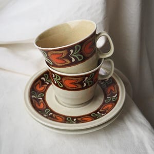 Könnte beinhalten: Zwei gestapelte beige Keramik-Teetassen mit passenden Untertassen. Jede Tasse und Untertasse hat ein sich wiederholendes Blumenmuster in Orange, Braun und Grün. Die Tassen haben einen gebogenen Griff und eine beige Innenseite.
