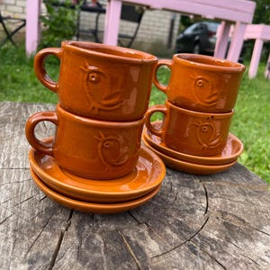 Peut inclure: Deux ensembles de tasses et soucoupes en céramique de couleur terre cuite. Chaque tasse présente un motif d'oiseau en relief sur le côté. Les tasses ont une anse arrondie et une finition lisse et brillante. Les soucoupes sont rondes.