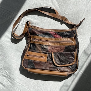 Könnte beinhalten: Eine mehrfarbige Umhängetasche aus Leder mit Patchwork-Design in Braun-, Schwarz- und Dunkelrottönen. Die Tasche verfügt über einen verstellbaren Schulterriemen, einen Reißverschluss oben, eine horizontale Reißverschlusstasche vorne und eine kleine Klappentasche.