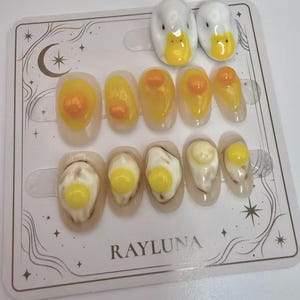 May include: A set of press-on nails with a fried egg design, featuring yellow yolks and white egg whites. Two duck-shaped nail charms are also included. The nails are displayed on a decorative card with the brand name "RAYLUNA".