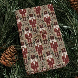 May include: A wrapped gift with a repeating pattern of red and brown nutcracker soldiers on a beige background. The gift is tied with twine and rests on a bed of green pine needles and pine cones, creating a festive Christmas scene.