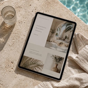 May include: A tablet displaying a website with the text "Intentional Beautiful Timeless" and images of interior design. A clear glass and a linen cloth are on a beige stone surface next to the tablet, with a pool of water in the background.
