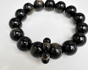 May include: A black beaded bracelet with a mix of large and small round beads. The larger beads have a reflective, polished surface. The bracelet is displayed on a white surface, showcasing its dark color and shine. The bracelet is a piece of jewelry.