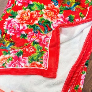 May include: A red blanket with a floral pattern featuring large pink and red flowers and green leaves. The blanket has a white, plush interior and a red border. Decorative birds are also visible in the design.