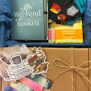 Pode incluir: Caixa de presente com tema de leitura. Inclui um caderno espiral turquesa com o texto "My Weekend is all Booked", uma caneta, meias com ilustrações de livros, chá, adesivos e marcadores. A caixa é amarrada com barbante.