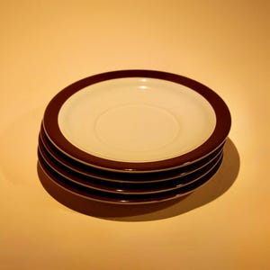 May include: A stack of five white ceramic plates with a dark brown rim. The plates are stacked on top of each other, with the top plate slightly angled. The plates are on a light orange background.