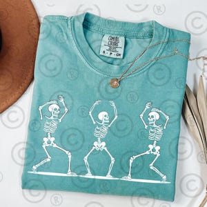 May include: Teal Comfort Colors t-shirt featuring a white graphic of three dancing skeletons. The skeletons are in different poses, with their arms raised. A gold necklace with a pendant is on the shirt.