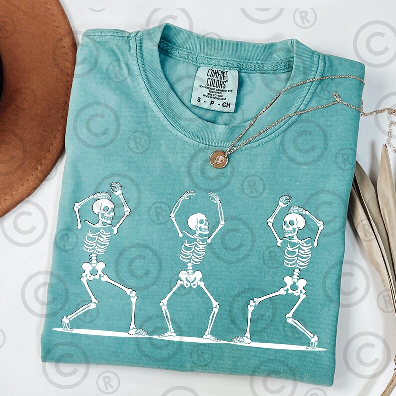 May include: Teal Comfort Colors t-shirt featuring a white graphic of three dancing skeletons. The skeletons are in different poses, with their arms raised. A gold necklace with a pendant is on the shirt.