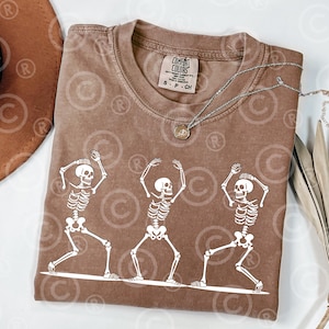 May include: A brown Comfort Colors t-shirt with a graphic of three white skeletons dancing. The shirt has a crew neck and short sleeves. A silver necklace with a pendant is draped across the shirt. The shirt is size S-P-CH.