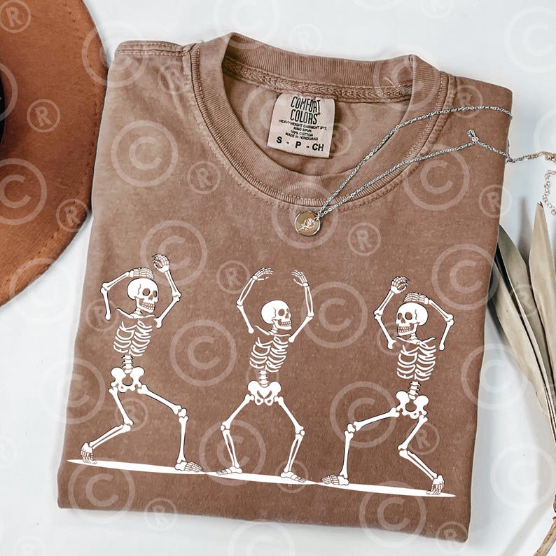May include: A brown Comfort Colors t-shirt with a graphic of three white skeletons dancing. The shirt has a crew neck and short sleeves. A silver necklace with a pendant is draped across the shirt. The shirt is size S-P-CH.