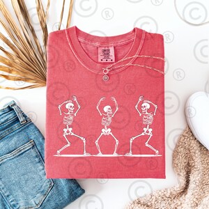 May include: A red Comfort Colors t-shirt with a white graphic of three skeletons in yoga poses. The shirt has a crew neck and short sleeves. A silver necklace with a sun pendant is draped across the shirt. The shirt is folded on a white surface.