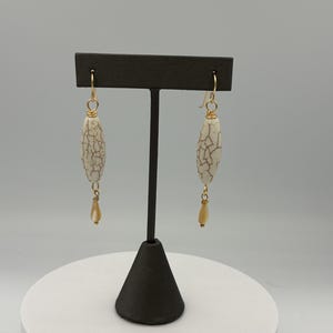 May include: A pair of dangle earrings displayed on a black stand. Each earring features a cream-colored, elongated bead with a cracked pattern, suspended from a gold-colored hook. A smaller, teardrop-shaped bead hangs below each main bead, adding a delicate touch.