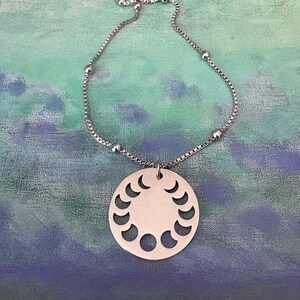 May include: Silver-toned necklace featuring a round pendant with crescent moon cutouts. The pendant hangs from a delicate chain with small, round beads. The background is a blend of blue and green hues.