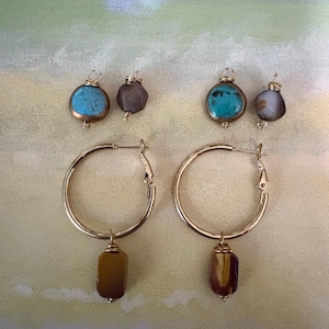 May include: Gold hoop earrings with dangling brown and yellow stone pendants. Two pairs of smaller earrings with turquoise and gray stones are also displayed. The jewelry is set against a backdrop of a painted beach scene.