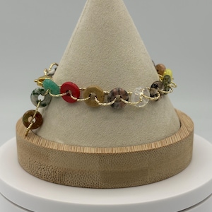 May include: A multi-coloured gemstone bracelet with beads in green, red, brown and clear, strung on gold-coloured wire. The bracelet is displayed on a beige cone-shaped stand with a wooden base, highlighting the design and craftsmanship.