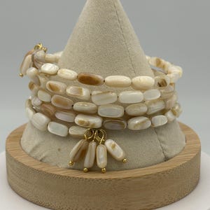 May include: A stack of ivory-colored beaded bracelets with gold accents. The bracelets are made of oval-shaped beads and are displayed on a beige cone-shaped stand. A cluster of dangling beads with gold caps hangs from the bracelets.