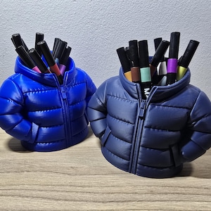 Op de afbeelding: Twee pennenbakjes in de vorm van pufferjacks, één in felblauw en de andere in een donkerdere blauwe kleur. Elke jas is gevuld met diverse gekleurde stiften. De jassen hebben een ritssluiting en staan op een houten oppervlak.