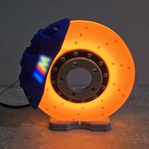 Pode incluir: Lâmpada decorativa em forma de disco e pinça de travão de carro. O disco é laranja com pequenos orifícios, e a pinça é azul com um logótipo multicolorido. A lâmpada emite um brilho laranja quente e assenta numa base cinzenta. Cerca de 25 cm de diâmetro.