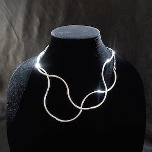Puede incluir: Un collar de plata con un diseño abstracto único. El collar está hecho de múltiples bucles interconectados, creando una forma fluida y orgánica.