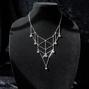 Pode incluir: Um colar geométrico prateado com detalhes de cristal transparente pendurado em um expositor de veludo preto.