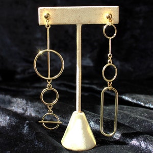 May include: Gold-toned geometric earrings displayed on a stand. One earring features a large circle with a vertical line, two smaller circles, and a horizontal line. The other earring has a series of circles and a long, rectangular shape.