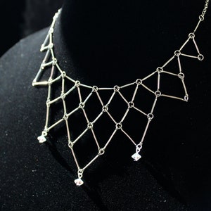 May include: A silver chain necklace with a geometric design and small crystal accents. The necklace is displayed on a black velvet mannequin.