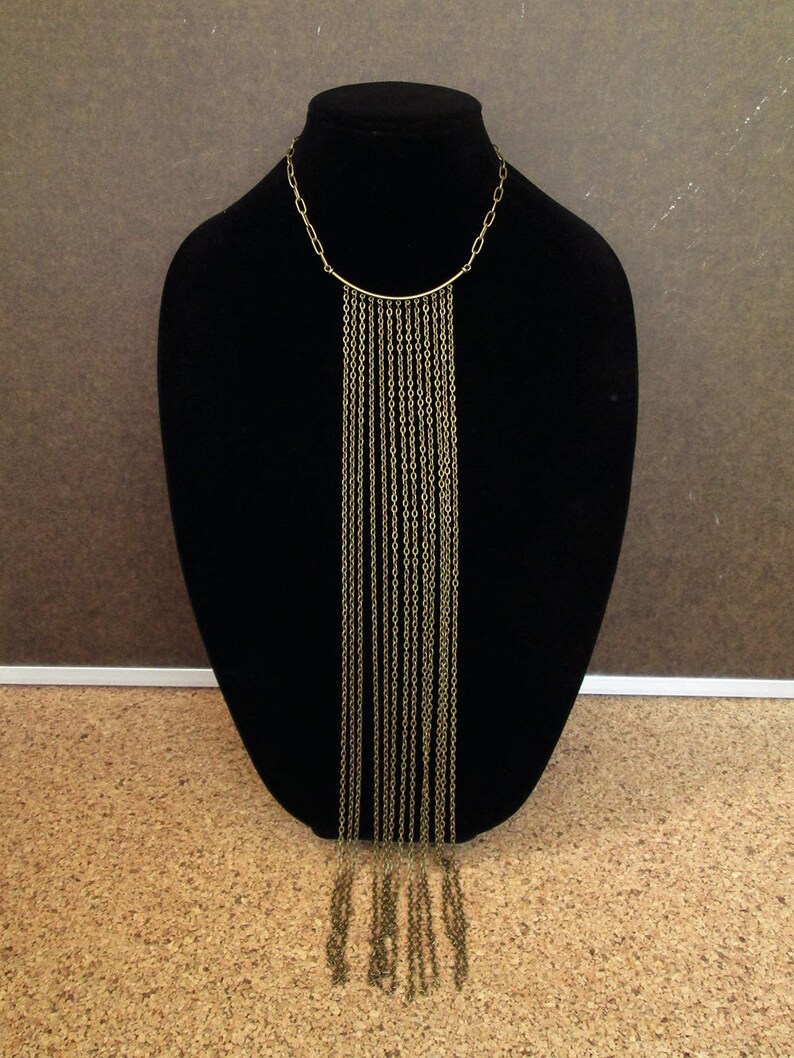 Puede incluir: Un collar largo de cadena dorada con una barra superior curva y m&uacute;ltiples hebras de cadena que cuelgan.