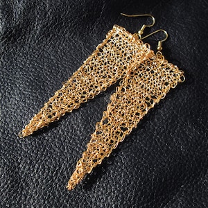 Puede incluir: Un par de pendientes de color dorado con forma triangular y un delicado diseño de ganchillo.