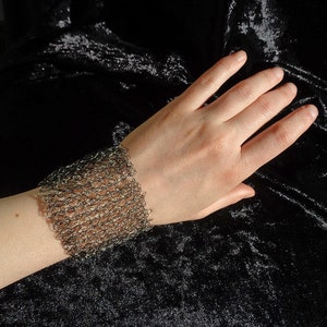 May include: A silver chainmail bracelet on a person's wrist. The bracelet is made of small, interconnected metal rings.