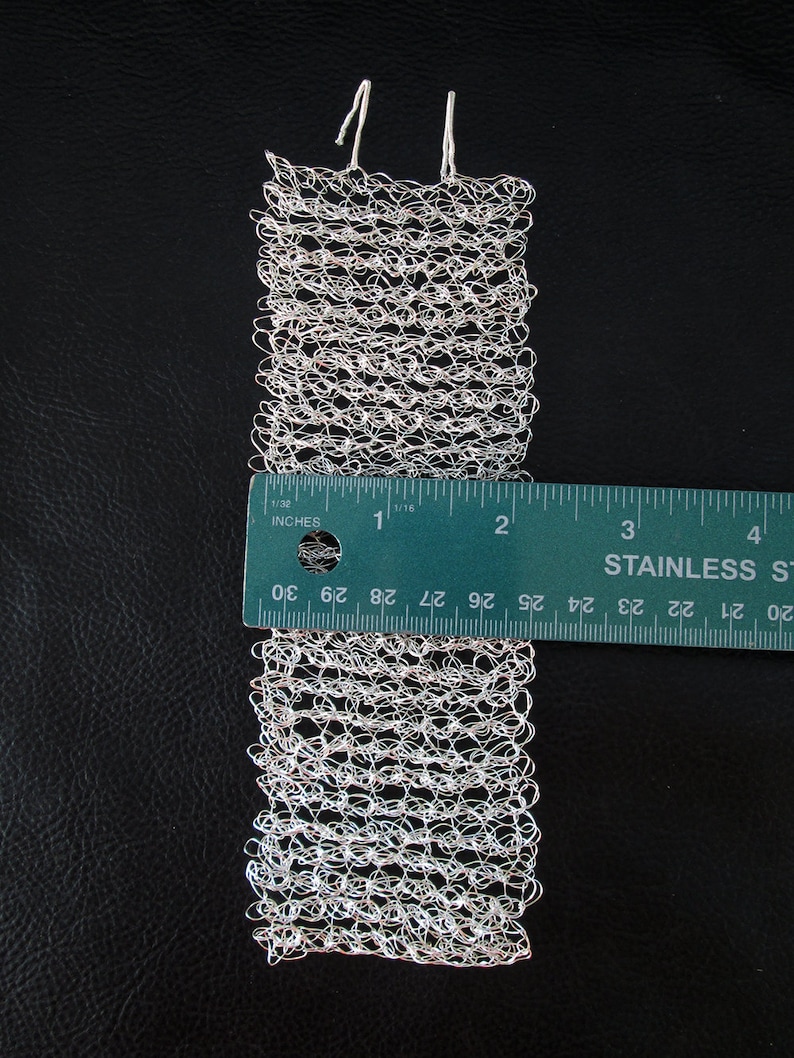 May include: A silver wire mesh bracelet with a textured, woven design. The bracelet is approximately 3 inches long and is measured against a ruler.