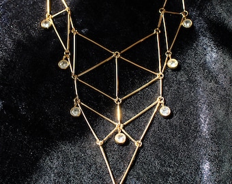 May include: A gold-toned necklace featuring a geometric, triangular wire frame structure. The design includes multiple interconnected metal bars forming a lattice pattern, accented with small, round, clear faceted stones hanging from the joints.
