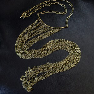 Puede incluir: Un collar de cadena dorada con una larga franja de cadenas m&aacute;s peque&ntilde;as que cae en cascada. El collar tiene una barra curva simple en la parte superior.