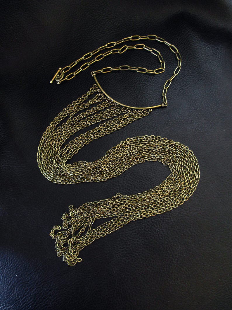 Puede incluir: Un collar de cadena dorada con una larga franja de cadenas m&aacute;s peque&ntilde;as que cae en cascada. El collar tiene una barra curva simple en la parte superior.