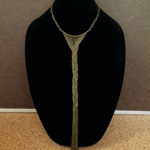 Puede incluir: Un collar largo de cadena dorada con una parte superior curva y un borla de cadenas que cuelga.
